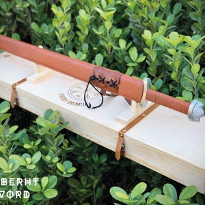 May include: A brown leather-wrapped sword with a silver pommel, resting in a wooden box with a leather strap and the inscription "Ulfberht Sword".