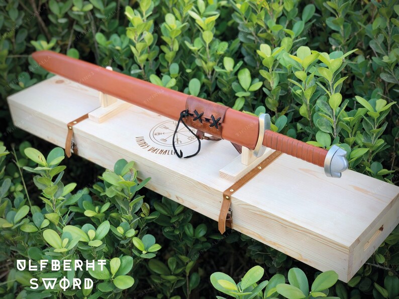 May include: A brown leather-wrapped sword with a silver pommel, resting in a wooden box with a leather strap and the inscription "Ulfberht Sword".