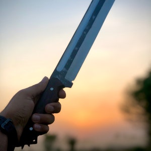 K&ouml;nnte beinhalten: Eine Person h&auml;lt ein gro&szlig;es Messer mit schwarzem Griff und silberner Klinge. Das Messer wird gegen einen Sonnenuntergangshimmel gehalten.