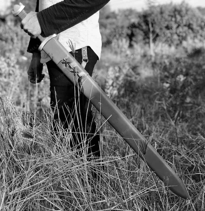 May include: A person holding a black and silver sword with a leather scabbard. The sword has a decorative design on the blade and a cross-shaped guard.