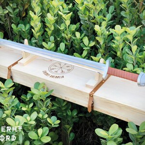 May include: A silver sword with a brown leather handle is displayed in a wooden box with a Viking symbol and the text "Odin's Valhalla".