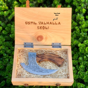 Könnte beinhalten: Holzkiste mit der Gravur "UNTIL VALHALLA SKOL!" und einer dekorativen Axt im Inneren. Die Axt hat einen Holzgriff und eine silberne Klinge mit einem komplizierten keltischen Knotenmuster. Die Kiste ist mit Stroh ausgekleidet.
