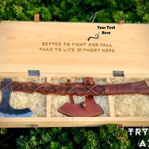 May include: A wooden box with a leather-wrapped axe inside. The box is open and the axe is visible. The box has a quote engraved on the lid: "Better to fight and fall than to live without hope". The axe has a leather handle with runes carved into it. The box is labeled "Frygve Axe".
