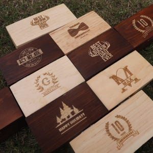 May include: A set of nine wooden boxes with various engraved designs. The designs include a bow tie, a deer head, a tree, a wreath, a heart, and the words "Best Buckin Dad Ever", "Happy Holidays", "Act My Age", "Thomas Groomman", "G Gonzalez", "M Max", and "10 Years Anniversary".