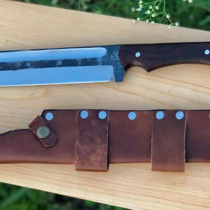K&ouml;nnte beinhalten: Ein gro&szlig;es, silbernes Messer mit einem braunen Holzgriff und einer Lederscheide mit silbernen Nieten. Das Messer hat eine lange, gebogene Klinge und eine scharfe Spitze.