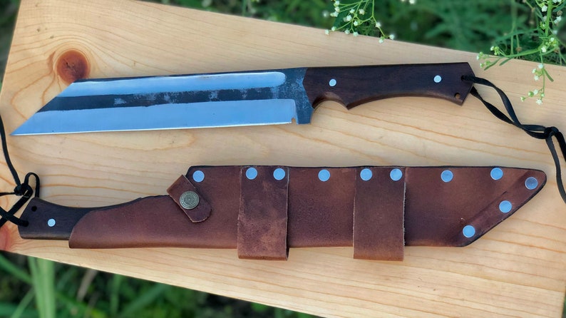 K&ouml;nnte beinhalten: Ein gro&szlig;es, silbernes Messer mit einem braunen Holzgriff und einer Lederscheide mit silbernen Nieten. Das Messer hat eine lange, gebogene Klinge und eine scharfe Spitze.