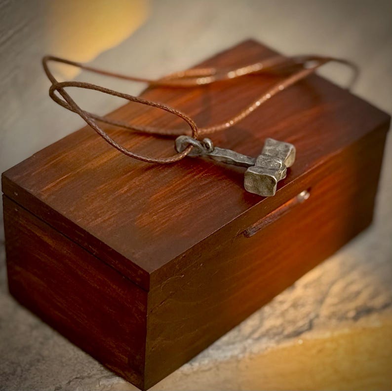 May include: A silver-toned hammer pendant necklace with a brown leather cord, displayed in a wooden box.