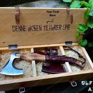 Puede incluir: Una caja de madera con una manija de cuero y un hacha de metal en su interior. La caja está grabada con el texto "DENNE ØKSSEN TILHØRER UWE" y "Your Text Here". El hacha tiene un mango de cuero y una cabeza de metal con un diseño de nudo vikingo.