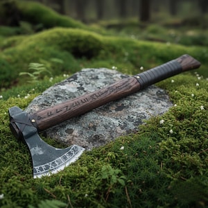 May include: A handcrafted axe with a dark wooden handle and a black blade. The handle is wrapped in black leather near the top. The blade has etched runes. The axe rests on a stone surrounded by green moss.