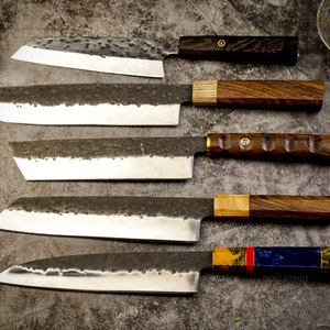 Pu&ograve; includere: Cinque coltelli da cucina diversi con manici in legno e motivi vari. I coltelli sono disposti verticalmente su una superficie grigia testurizzata.