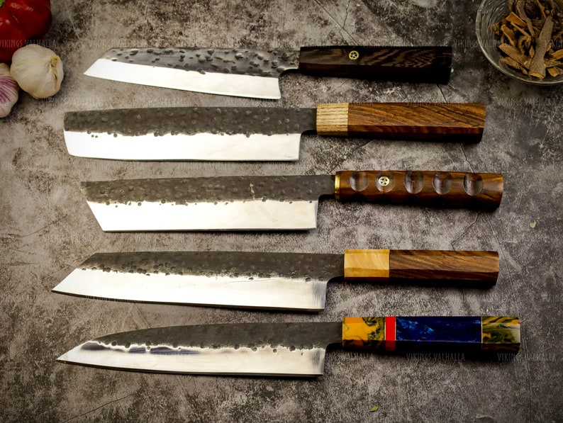 Pu&ograve; includere: Cinque coltelli da cucina diversi con manici in legno e motivi vari. I coltelli sono disposti verticalmente su una superficie grigia testurizzata.