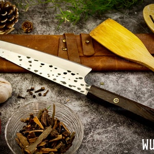 Pu&ograve; includere: Un coltello da cucina in acciaio inossidabile con manico in legno nero e finitura martellata. Il coltello &egrave; appoggiato su una superficie in pelle marrone con un cucchiaio di legno e una spatola. Il coltello ha la scritta "WUNJO" stampata sul manico.