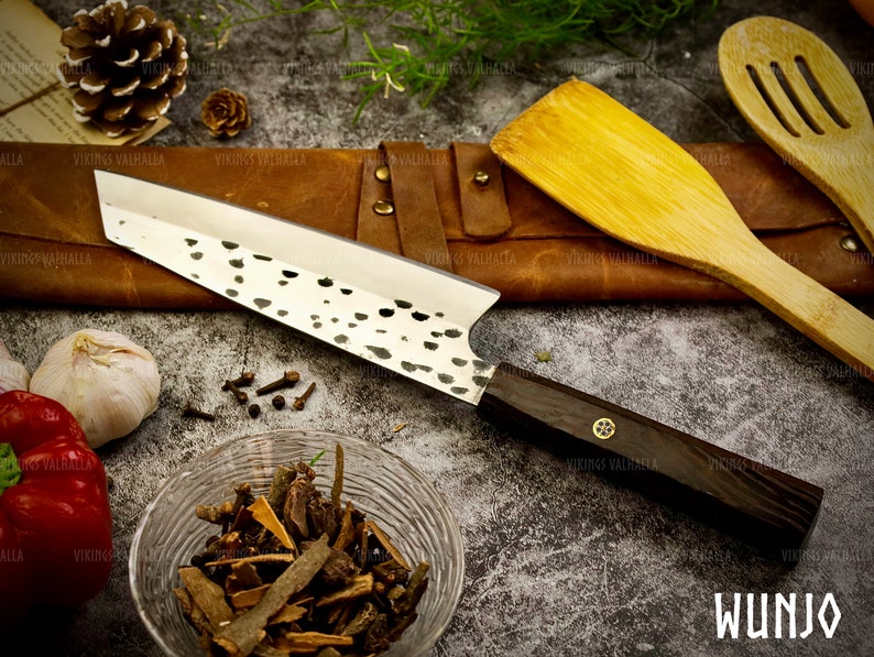 Pu&ograve; includere: Un coltello da cucina in acciaio inossidabile con manico in legno nero e finitura martellata. Il coltello &egrave; appoggiato su una superficie in pelle marrone con un cucchiaio di legno e una spatola. Il coltello ha la scritta "WUNJO" stampata sul manico.