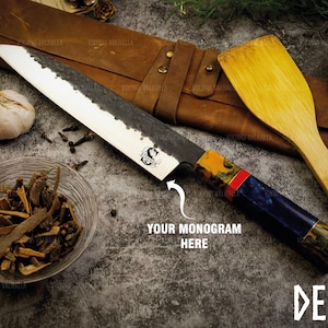 May include: A black and blue Japanese deba knife with a wooden handle and a red and yellow pattern. The knife is on a brown leather roll with the text "VIKINGS VALHALLA" printed on it. The knife is next to a wooden spatula and a wooden spoon. The text "DEBA" is printed on the bottom right corner of the image. The text "YOUR MONOGRAM HERE" is printed in the middle of the image.