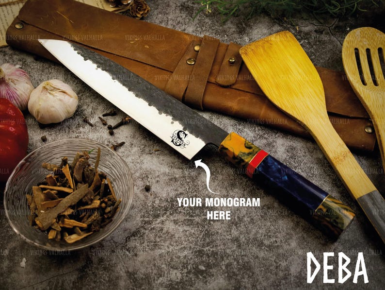 Pu&ograve; includere: Un coltello giapponese deba nero e blu con manico in legno e un motivo rosso e giallo. Il coltello &egrave; su un rotolo di cuoio marrone con il testo "VIKINGS VALHALLA" stampato su di esso. Il coltello &egrave; accanto a una spatola di legno e un cucchiaio di legno. Il testo "DEBA" &egrave; stampato nell'angolo in basso a destra dell'immagine. Il testo "YOUR MONOGRAM HERE" &egrave; stampato al centro dell'immagine.