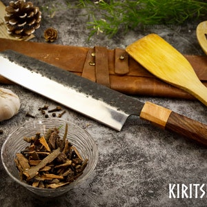 Pu&ograve; includere: Un coltello Kiritsuke con manico in legno e fodero in pelle. Il coltello &egrave; su una superficie grigia con spezie e verdure.