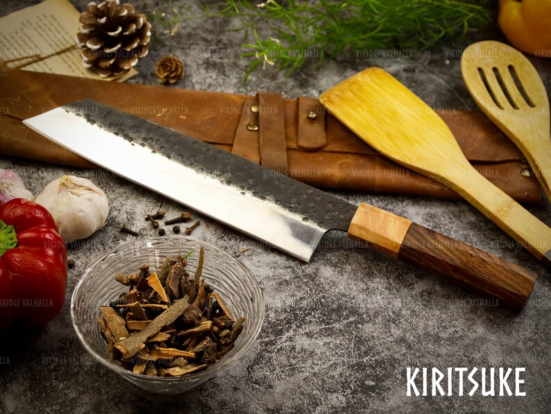 Pu&ograve; includere: Un coltello Kiritsuke con manico in legno e fodero in pelle. Il coltello &egrave; su una superficie grigia con spezie e verdure.