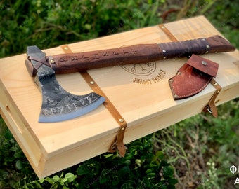 Può includere: Un'ascia vichinga forgiata a mano con manico in legno e avvolgimento in pelle. L'ascia è incisa con rune e la parola "AXE". È esposta in una scatola di legno con un fodero in pelle.