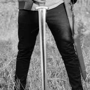 May include: A person holding a silver sword with a black leather scabbard. The sword has a wooden handle and a long, sharp blade. The person is wearing black trousers and a jacket.