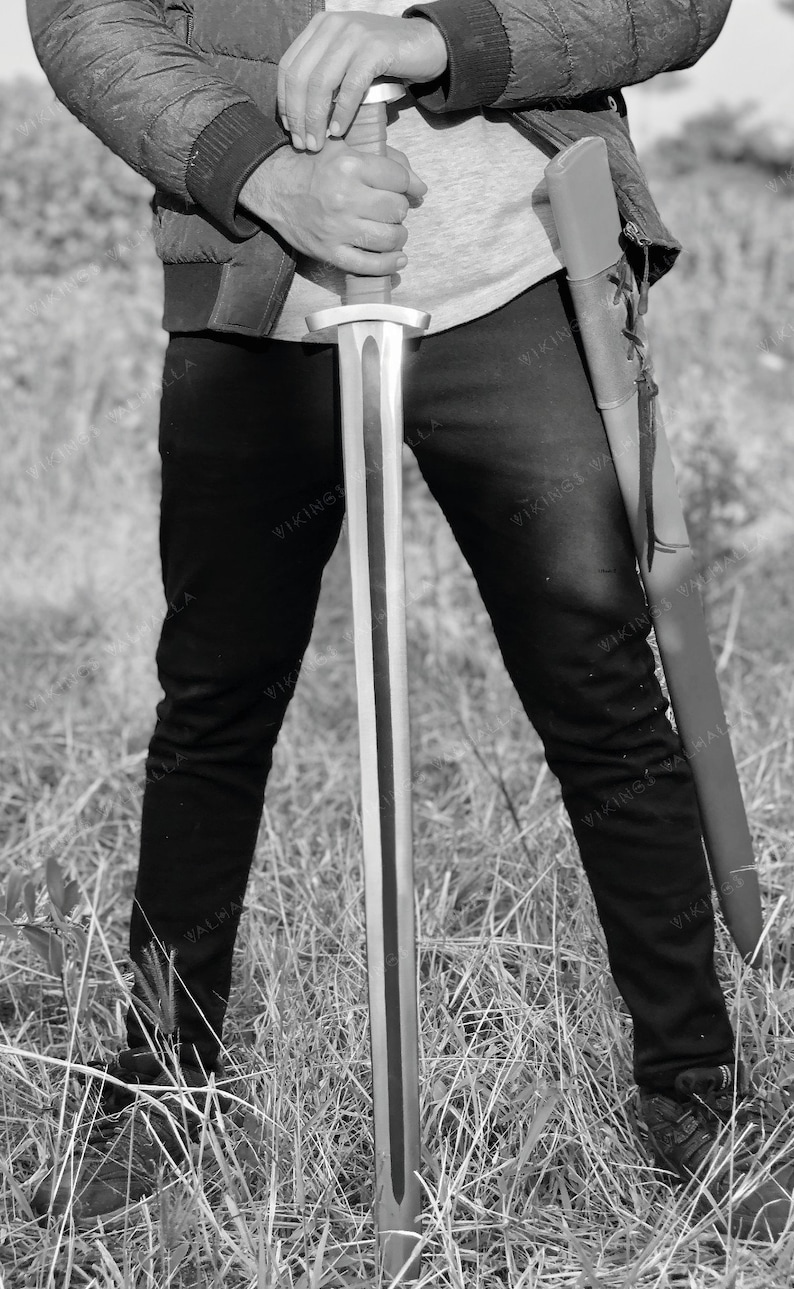 May include: A person holding a silver sword with a black leather scabbard. The sword has a wooden handle and a long, sharp blade. The person is wearing black trousers and a jacket.