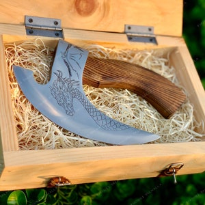 Könnte beinhalten: Eine dekorative Axt mit einem Holzstiel und einer gebogenen Klinge. Die Klinge ist mit einem Drachenmuster graviert. Die Axt wird in einer Holzkiste mit strohähnlichem Verpackungsmaterial präsentiert.
