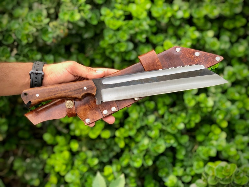 K&ouml;nnte beinhalten: Eine braune Lederscheide mit einem silbernen Messer mit einem Holzgriff. Das Messer ist scharf und hat eine gebogene Klinge.