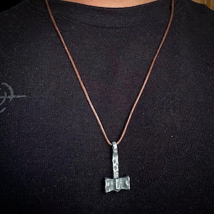 May include: A silver-colored hammered metal pendant in the shape of a Mjolnir, the Norse god Thor's hammer, hangs from a brown leather cord.