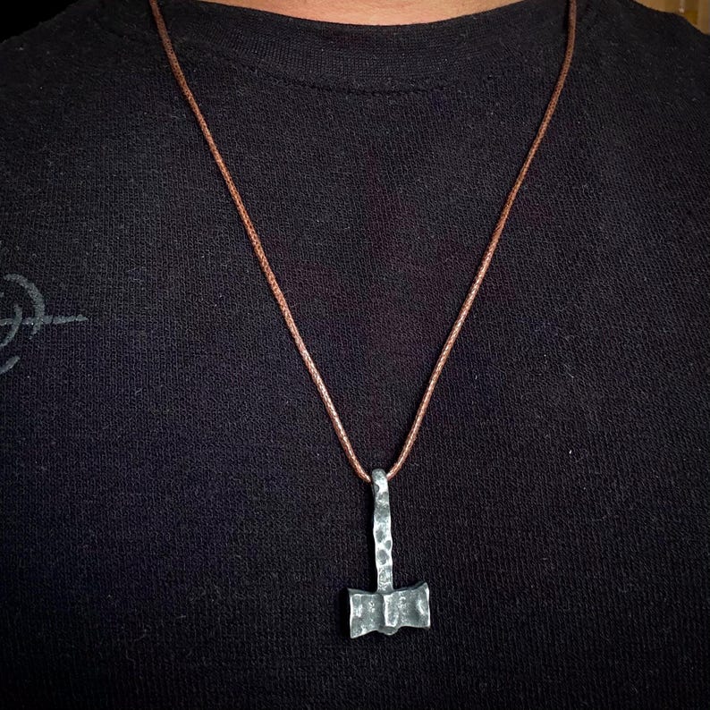 May include: A silver-colored hammered metal pendant in the shape of a Mjolnir, the Norse god Thor's hammer, hangs from a brown leather cord.