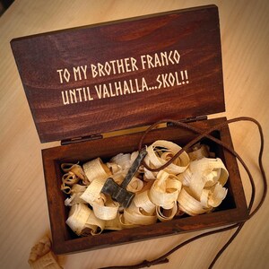 May include: A wooden box with a hinged lid, engraved with the message "To my brother Franco Until Valhalla... Skol!!". The box is open, revealing a small axe pendant on a brown cord, nestled in wood shavings.