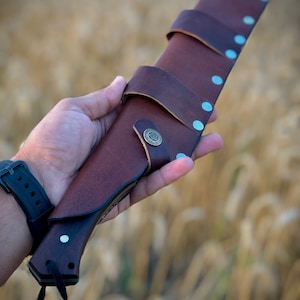 K&ouml;nnte beinhalten: Ein braunes Lederscheide mit einem Metallknopf und Nieten, in dem ein Messer mit einem Holzgriff steckt. Die Scheide wird in einer Hand vor einem verschwommenen Hintergrund aus goldenem Weizen gehalten.