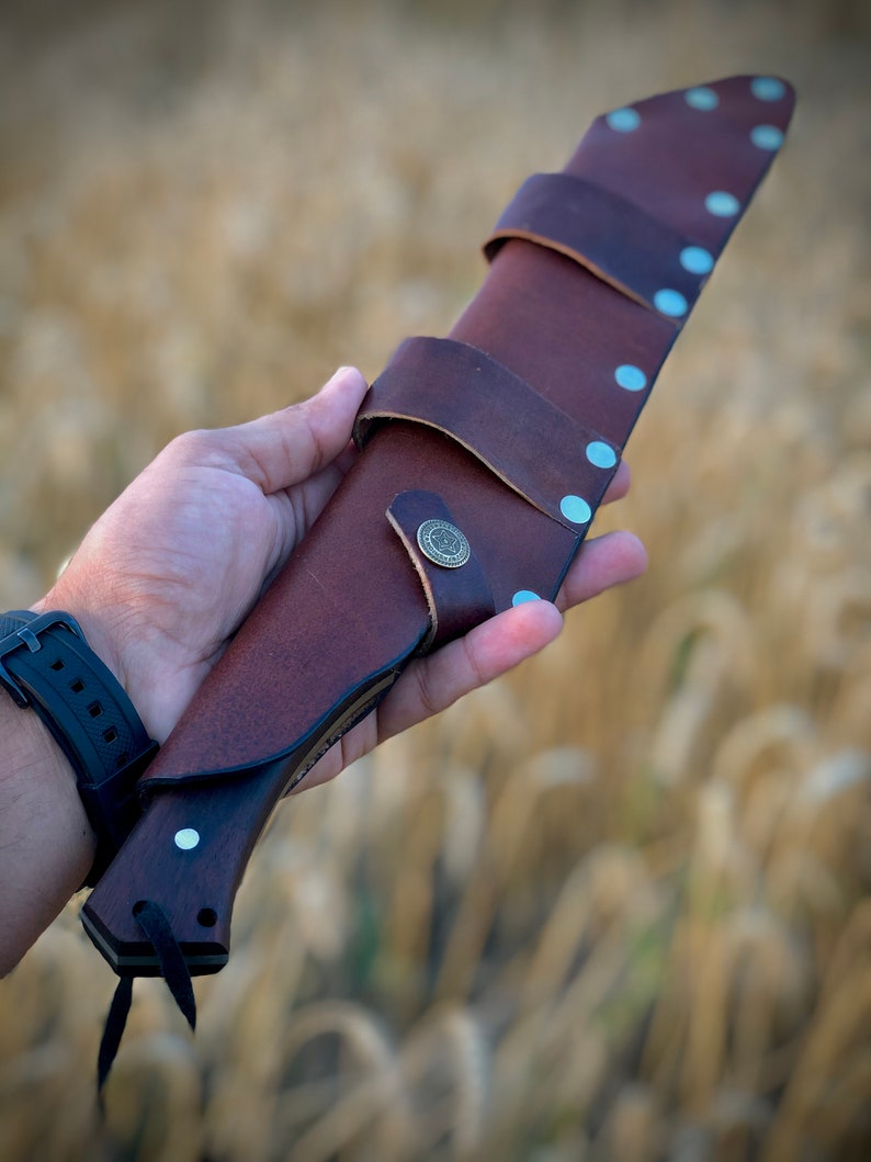 K&ouml;nnte beinhalten: Ein braunes Lederscheide mit einem Metallknopf und Nieten, in dem ein Messer mit einem Holzgriff steckt. Die Scheide wird in einer Hand vor einem verschwommenen Hintergrund aus goldenem Weizen gehalten.