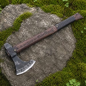May include: A hand axe with a dark metal head featuring etched runic symbols along the curved blade edge. The handle is crafted from dark wood with a textured grain, displaying carved runic inscriptions and a black leather-wrapped grip section.