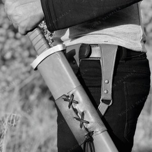 May include: A brown leather scabbard with a black strap and a white metal tip. The scabbard is decorated with a criss-cross pattern of black leather straps.
