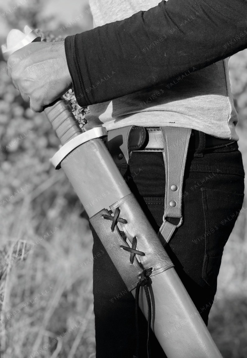 May include: A brown leather scabbard with a black strap and a white metal tip. The scabbard is decorated with a criss-cross pattern of black leather straps.