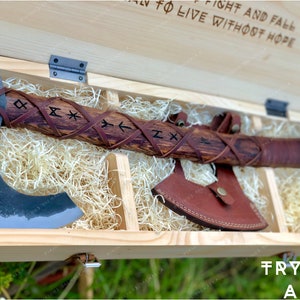 Könnte beinhalten: Eine Holzkiste mit einer lederumwickelten Axt darin. Die Axt hat einen Holzstiel mit eingegrabenen Runen. Die Kiste ist mit dem Text "Fight and fall to live without hope" und "Trygve Axe" graviert.