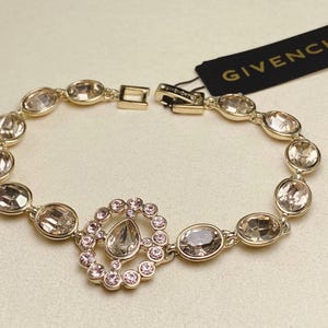 May include: A gold-toned bracelet with oval-shaped, clear gemstones. The bracelet features a central design of a teardrop-shaped stone surrounded by smaller pink stones. A black tag with the word "GIVENCHY" is attached.