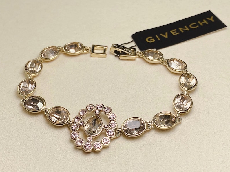 May include: A gold-toned bracelet with oval-shaped, clear gemstones. The bracelet features a central design of a teardrop-shaped stone surrounded by smaller pink stones. A black tag with the word "GIVENCHY" is attached.