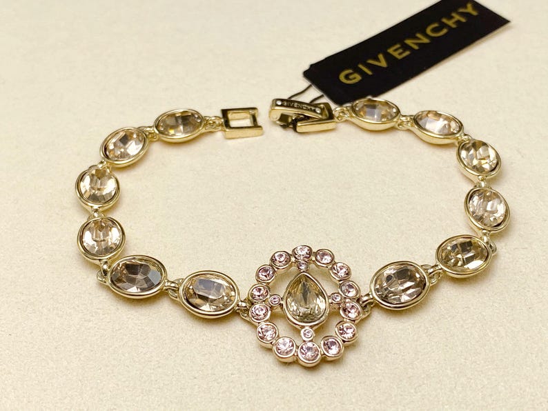 May include: Gold-toned bracelet with oval-shaped crystals and a central circular design with smaller pink crystals. The bracelet has a clasp closure and a Givenchy tag.