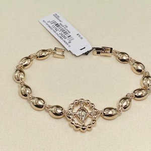 May include: Gold-toned bracelet featuring oval-shaped links and a decorative centerpiece. The bracelet has a clasp closure and a tag attached. The tag includes text that reads "MADE IN CHINA" and "NOT INTENDED FOR CHILDREN UNDER 15".