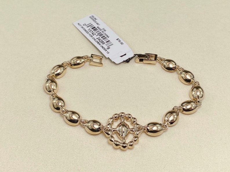 May include: Gold-toned bracelet featuring oval-shaped links and a decorative centerpiece. The bracelet has a clasp closure and a tag attached. The tag includes text that reads "MADE IN CHINA" and "NOT INTENDED FOR CHILDREN UNDER 15".