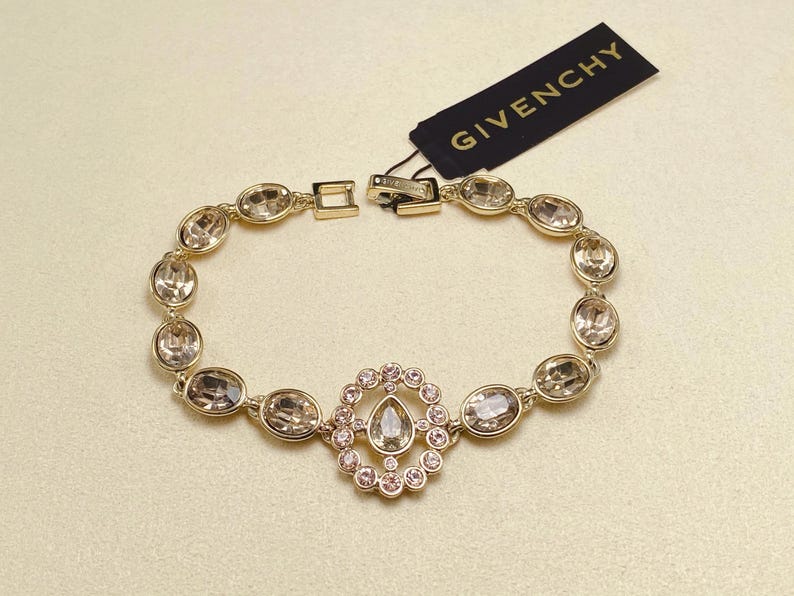 May include: Gold-toned bracelet with oval and round clear crystals. A central design features a teardrop-shaped crystal surrounded by smaller pink crystals. The tag reads "GIVENCHY".