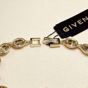 May include: Gold-toned bracelet featuring oval-shaped, clear gemstones. The bracelet is connected with a clasp and has a Givenchy tag attached. The gemstones are set in a gold metal frame, creating a luxurious look.