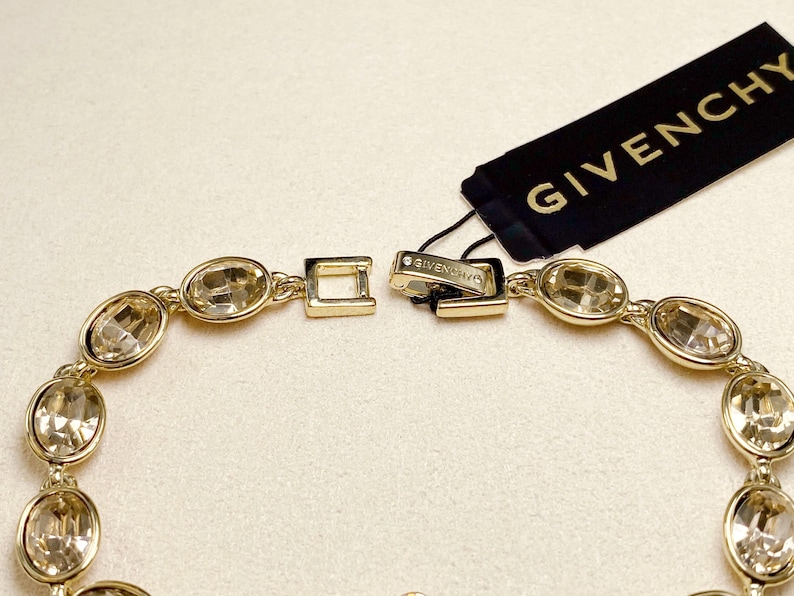 May include: Gold-toned bracelet featuring oval-shaped, clear gemstones. The bracelet is connected with a clasp and has a Givenchy tag attached. The gemstones are set in a gold metal frame, creating a luxurious look.