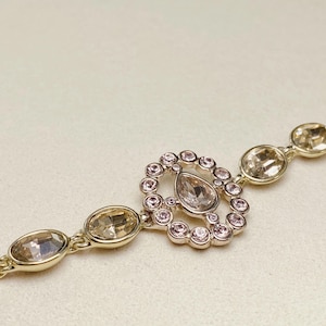 May include: A gold-toned bracelet featuring a central pendant with a teardrop-shaped crystal surrounded by small pink crystals. The bracelet has oval-shaped crystals and a delicate chain.