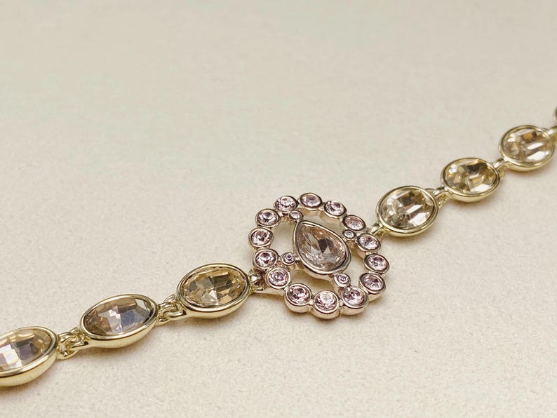 May include: A gold-toned bracelet featuring a central pendant with a teardrop-shaped crystal surrounded by small pink crystals. The bracelet has oval-shaped crystals and a delicate chain.