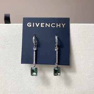 May include: A pair of silver-toned drop earrings with a rectangular, olive-green gemstone at the bottom. The earrings are attached to a dark blue card with the word "GIVENCHY" in gold lettering.