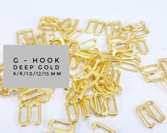 May include: A pile of gold-coloured metal G-hook bra clasps in various sizes, 6/8/10/12/15 mm.  G-HOOK DEEP GOLD 6/8/10/12/15 MM is written on a white label.
