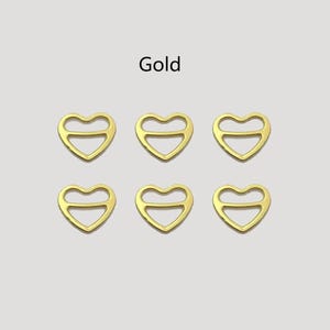 Peut inclure: Six boucles en métal dorées en forme de cœur. Chaque boucle a un contour de cœur avec une barre horizontale en haut. Le mot "Gold" est imprimé au-dessus des boucles.