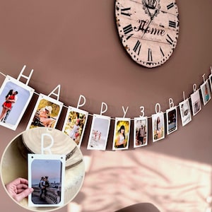 Może przedstawiać: Biały wiszący baner z napisem HAPPY BIRTHDAY z wbudowanymi klipsami do mocowania prostokątnych zdjęć. Każda litera jest wycięta z białego materiału i służy jako klips do zdjęcia, tworząc możliwość personalizacji dekoracji.