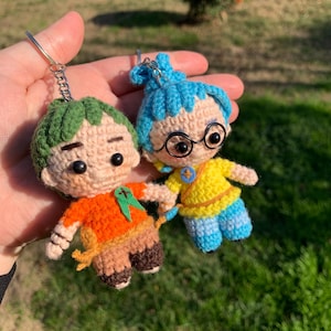 Può includere: Due figure di portachiavi all'uncinetto, una con i capelli verdi e una camicia arancione, l'altra con i capelli blu e una camicia gialla. Entrambe le figure hanno gli occhi marroni e si tengono per mano.